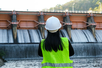 Rear view of female engineer in green vest and helmet standing outside against background of dam with hydroelectric power plant and irrigation. - obrazy, fototapety, plakaty