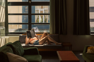 young woman in summer clothes lying on the couch reading a book in the warm daylight under a big window