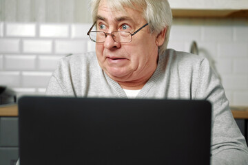 Senior grey haired man wearing glasses sitting at laptop and making fish face with lips. Crazy and...