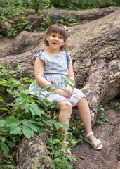Naklejka na ściany i meble A cute little girl sits on huge tree roots, walks in the fresh air in summer, the child is wearing a summer dress. Summer sunny day. Children's holidays. 