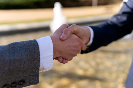 Anonymous businessmen in formal suits shaking hands while standing near blurred fountain during meeting