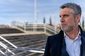 Confident bearded senior businessman in formal wear looking away while standing on stairs and thinking about project during walk
