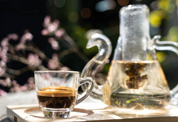 Espresso shot coffee in clear small cup with blur pink flower in background in the morning , there is blur  teapot place beside on the table