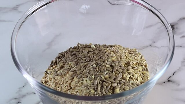 A female hands puts a ingredients of granola to a glass mixing bowl on a marble background. Healthy breakfast from oat sereals, nuts, seeds and eco sweetener dates syrup. Close up