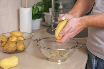 A man peels potatoes with a vegetable peeler in a bright kitchen. Preparation of potatoes for cooking. Cleaning peel from pesticides. A man cooks dinner or lunch at home for family.