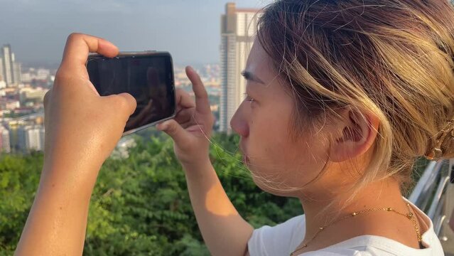 Beautiful young asian woman hold phone in hands take picture and video of landscape panoramic city top view outdoor sunny day, Pattaya, Thailand. Tourism and travel concept