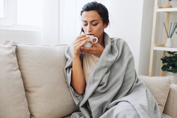 Woman cold headache with a cup of tea sits on a sofa in a plaid, treatment for flu, allergies and covid-19