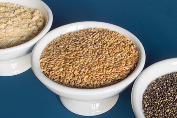 Close-up of a small plate full of golden flax seeds surrounded by two bowls of chia seeds and flax powder on a blue studio background. Healthy seeds to eat superfood.