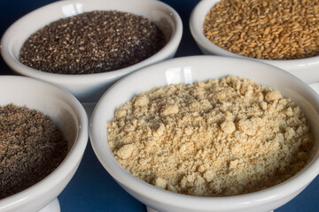 Close-up of a small plate filled with powdered golden flax seeds surrounded by small bowls of whole chia and flax seeds. Healthy seeds to eat superfoods.
