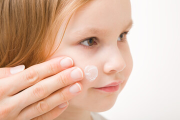 Young adult mother finger applying white moisturizing cream on toddler girl cheek. Daily care about baby face skin. Closeup. Side view. Isolated on light gray background.