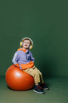 Stylish little boy in headphones sits on orange swiss ball on green background