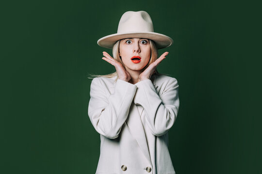 Suprised stylish blond hair woman in white blazer and hat on green background