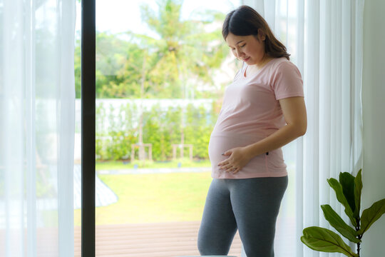 Pregnancy, people rest and expectation concept - Asian smiling happy pregnant woman standing near window and touching her belly at home.