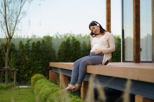 Portrait of pregnant Happy Asian woman sitting on terrace at home and using smart phone for listening music in headphones.