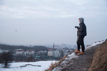 osoba w kapturze stojąca na wzgórzu z aparatem - obrazy, fototapety, plakaty