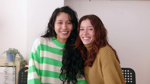 Young adult women smiling at camera while sitting at living room table. Female friendship concept with two cheerful latin american females together.