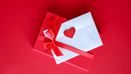 Romantic love letter in white envelope with Valentines heart and gift box with red ribbon on red background. Flat lay, top view, copy space. Happy Valentine's Day concept