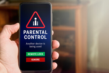 Male hand holding mobile phone with parental control warning on the screen: "Another device is being used". Buttons with options to remote lock or ignore. Children safety and care.