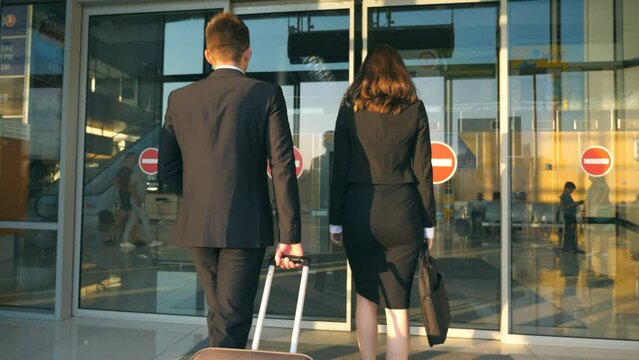 Business man and woman going to the airport with their luggage. Young businessman carrying suitcase on wheels and walking with his female colleague to terminal hall. Trip or travel concept Slow motion