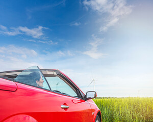 Fototapeta premium Woman in a red car against the background of a field and blue sky outside the city.