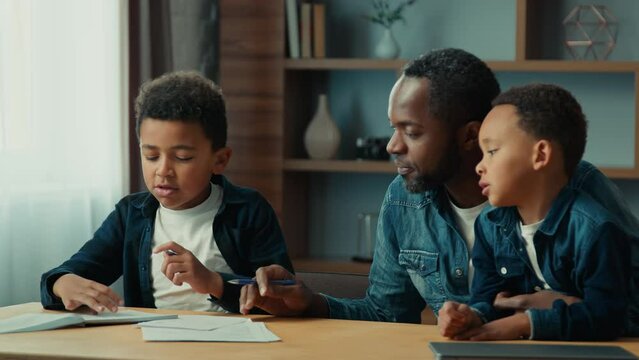 African American father with two little sons boys children sitting at table dad teach help pupil schoolboy boy son write writing lesson class helping with homework studies home learning homeschooling