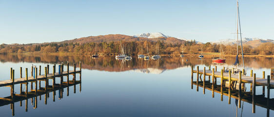 Ambleside Pier with Warm light bathing the boats on Lake Windermere with perfect reflections. Snow Capped mountains in the background.  Lake District Panorama. High Megapixel banner image