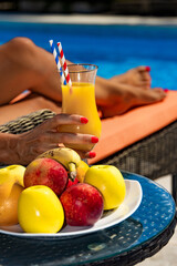 Une femme boit un cocktail de fruit devant une  piscine et une belle corbeille de fruits frais 