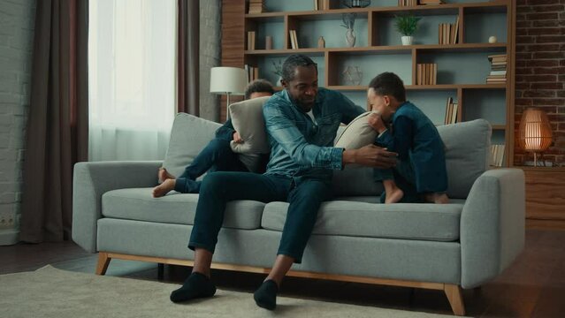 African American ethnic man father dad sitting at couch at home little noise active boys children sons play game on sofa hyperactive kids fighting pillows having fun fight difficulties of upbringing