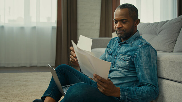 Mature african american man sitting in living room reading documents ethnic businessman freelancer working with papers analyzes finance checking report entering data in laptop work corporate paperwork