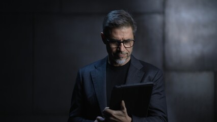 Portrait of mature businessman, using tablet computer. Entrepreneur standing in front of dark loft...