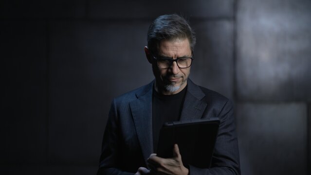 Portrait of mature businessman, using tablet computer. Entrepreneur standing in front of dark loft wall. Older, middle aged, mid adult, man in his 40s or 50s in jacket, business casual.