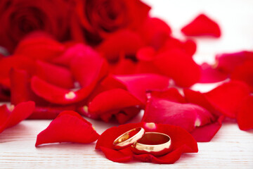 Wedding rings and many rose petals and a bouquet of red roses on white wooden desk