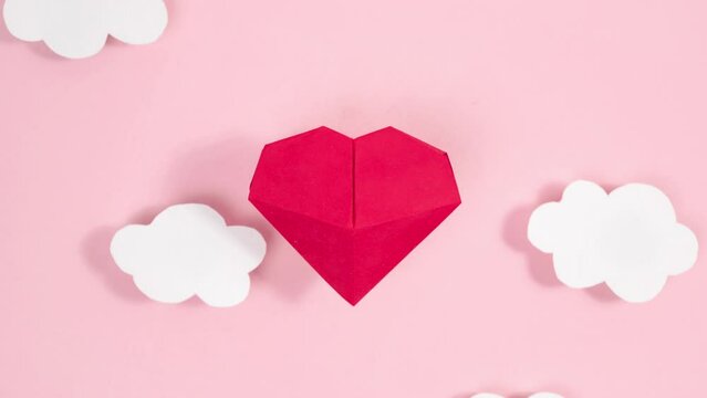 4k Volumetric origami heart swaying flies between white clouds. Greeting card. Pink background. Symbol of love. Concept of valentine's holiday, wedding and other occasions. Loop stop motion animation.