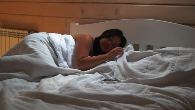 Girl wakes up in the morning and turns off the alarm clock on the phone in bed with a white blanket in a wooden house