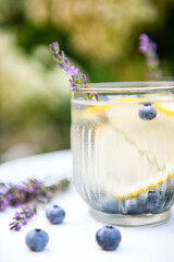 Close up of sweaty glass with cold delicious homemade lemonade made of fresh blueberries, lemon and lavender on white table on backyard in summer day and blooming hydrangea flowers on background.