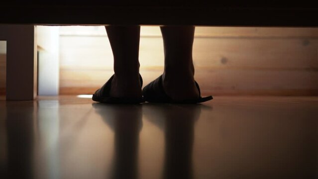 Women's legs get out of bed, put on slippers and leave against the backdrop of a wooden house under the rays of the morning sun