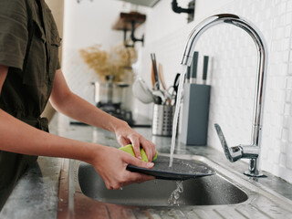 A woman with her hands without gloves washes dishes with a sponge, household chores, no dishwasher, high water consumption. Close-up of hands