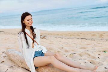 Happy tanned woman smiling with teeth in white swimsuit shirt and denim shorts sits on the sand by the ocean with wet hair after swimming, sunset light and pink clouds in Bali