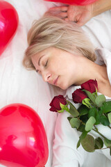 a cute young woman  with red roses is lying on white bed.