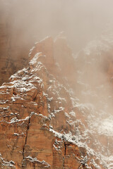Low clouds partially hide the red sandstone fins laced with snow on a steep mountain slope in Zion National park, Utah, USA in the middle of winter.