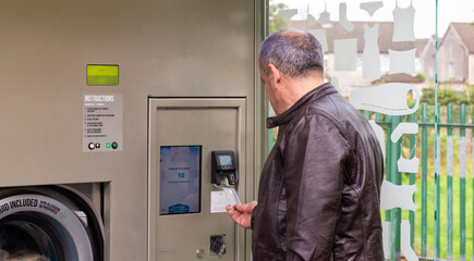 senior male, wearing jeans and brown jacket, using self-service laundry, inserting dirty clothes and using card payment, horizontal image, copy space