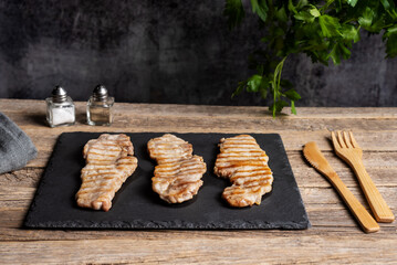 Iberico pork loin steaks made on the grill on a black stone plate with wooden cutlery on a rustic old wooden table, next to salt, pepper and parsley leaves. Close-up.