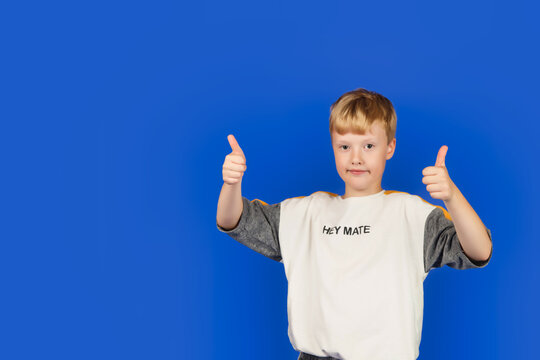 Little boy in white tshirt with HEY MATE text thumbs up hands posing on blue isolated background, looking at camera. Stylish smiling guy standing in studio. Advertising concept. Copy text space