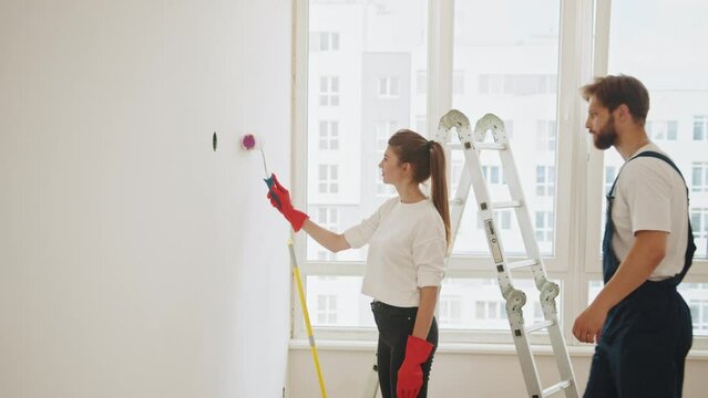 Couple Decorating Room In New Home Painting Wall Together. Cheerful couple young man and woman, painting white wall with roller during renovation in new apartment. Couple is repairing.