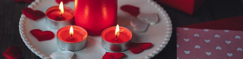 Saint Valentine's Day celebration. Red burning candles, hearts, gift box, postcard on dark wooden background. Happy holiday. Table decor for festive dinner, romantic atmosphere. Banner