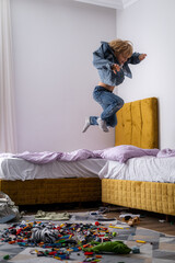 boy having fun, jumping on bed in messy childrens room, kid playing among the many toys at home, a...