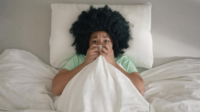 Frightened African American woman scared of ghosts on bed hiding under blanket at early morning. Top view on woman of color lying in bed and covering face by blanket to not see scary things at home 4K