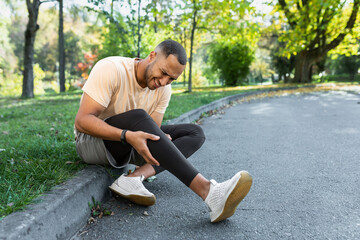 Man injured leg while jogging, african american man sitting on ground, massaging sore muscle with hands, upset and injured runner. - obrazy, fototapety, plakaty