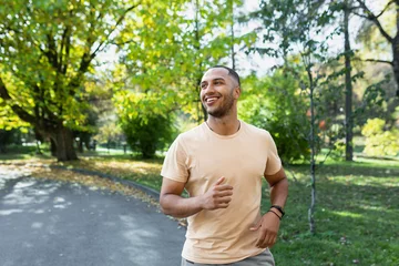 Zelfklevend Fotobehang Cheerful and successful hispanic man jogging in the park, man running on a sunny day, smiling and happy having an outdoor activity. © Liubomir
