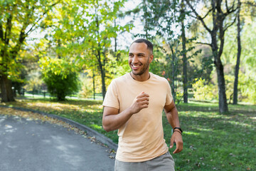 Naklejka na ściany i meble Cheerful and successful hispanic man jogging in the park, man running on a sunny day, smiling and happy having an outdoor activity.